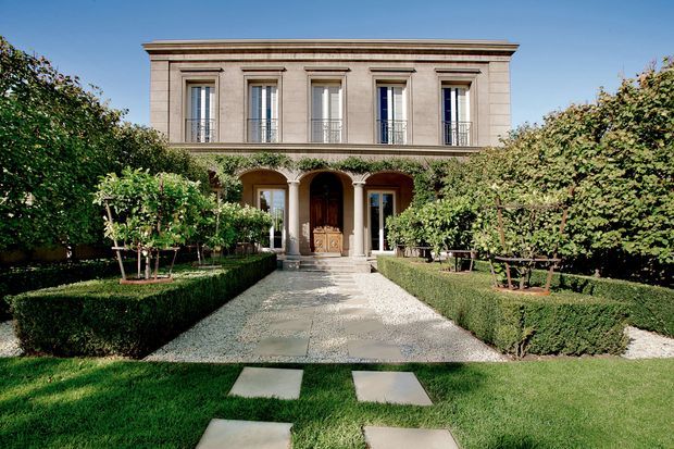 Formal two-storey classical residence viewed from a landscaped front garden. The symmetrical rendered facade has tall recessed windows with decorative surrounds, a projecting cornice, arched ground-floor openings, slender columns, and a central timber entry door. Clipped hedges, espaliered greenery, gravel paths, and large stone stepping slabs create a structured garden approach.