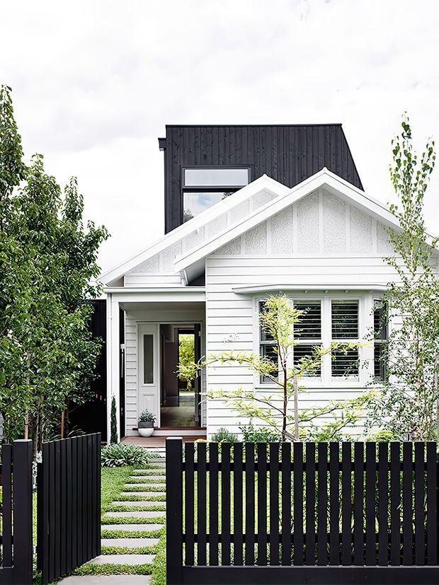 Front exterior view of a compact residential house with a white weatherboard facade, gabled roof forms, decorative gable infill, and a contrasting tall black vertically clad upper volume. A black vertical-slat fence, paved stepping-stone path, covered entry, timber-framed windows, and established front planting complete the contemporary streetscape.