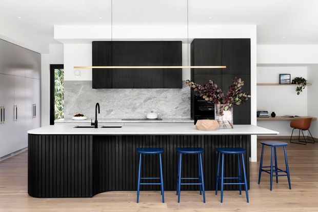 Contemporary open-plan kitchen with a large white island, dark fluted cabinetry, pale stone splashback, integrated appliances, black tapware, and blue bar stools. The space opens toward a dining or living area with timber flooring and minimal shelving.