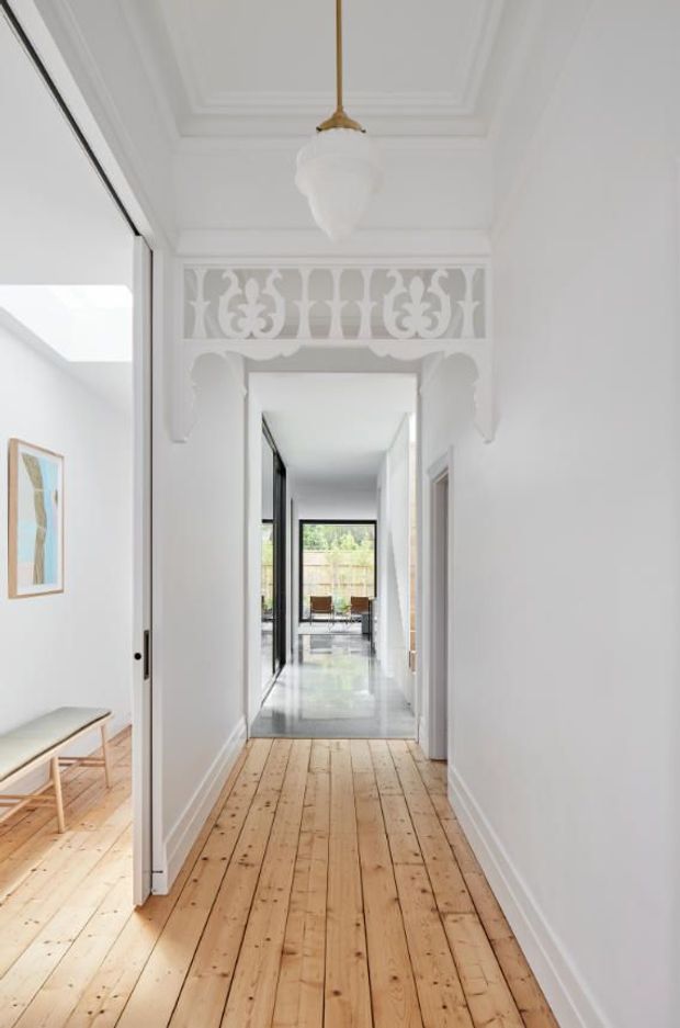 Bright, narrow residential hallway with original timber floorboards, white walls, decorative moulded ceiling, and ornate white plaster detailing above the doorway. A sculptural pendant light hangs centrally. The corridor leads toward a glazed living area at the rear, with a partially visible bright room and bench to the left.