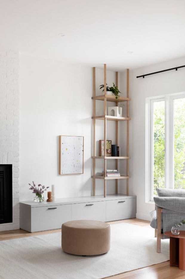 Bright, calm living room with a white-painted brick fireplace wall, low white built-in media cabinetry, and an open timber shelving unit displaying books and decor. A grey upholstered armchair sits beside tall white-framed windows, with a rounded taupe ottoman on a pale rug over timber flooring. Minimal artwork, greenery, and simple black curtain hardware complete the space.