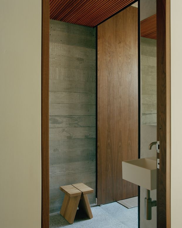 A compact, architecturally detailed bathroom viewed through a timber-framed opening. The space features a grey textured concrete or cement wall, a warm timber slatted ceiling, a tall timber door or wall panel with black metal framing, and a floating white rectangular basin with exposed plumbing. A small timber stool sits on a light speckled floor, and a mirror reflects the concrete wall and ceiling.
