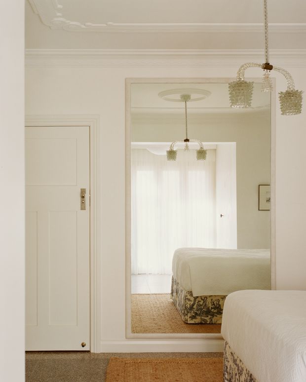 A softly lit, elegant bedroom interior dominated by warm white tones. The image shows a paneled white door, ornate plaster ceiling mouldings, a large full-length mirror, a crystal pendant light, sheer curtains, and the edge of a neatly made bed with patterned bed skirt. The mirror reflects another bed and the bright window, reinforcing the bedroom setting.