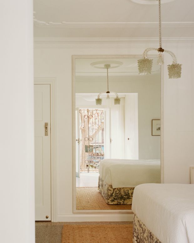 A softly lit, elegant bedroom interior featuring two white-covered beds with patterned bed skirts, a large full-height wall mirror, ornate ceiling cornices and rosettes, and delicate hanging pendant lights. The mirror reflects a second bed, a doorway, and a bright glazed exterior opening. The palette is predominantly warm white, cream, and beige with traditional decorative detailing.
