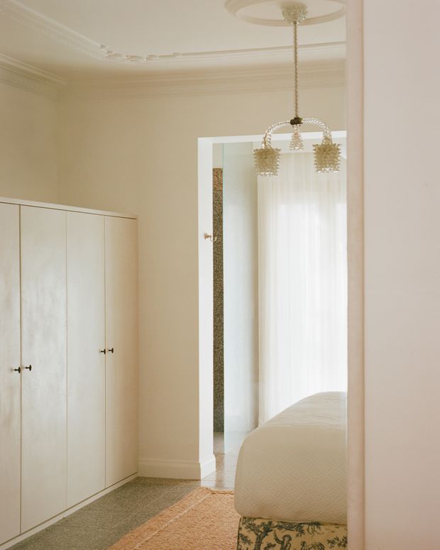 A softly lit, refined bedroom interior viewed from an adjoining space. Cream built-in wardrobe cabinetry lines the left wall, while an open doorway reveals a bed with a floral upholstered base, sheer curtains, and a decorative crystal pendant. The room features ornate plaster ceiling detailing, a ceiling medallion, warm neutral finishes, and a textured area rug.