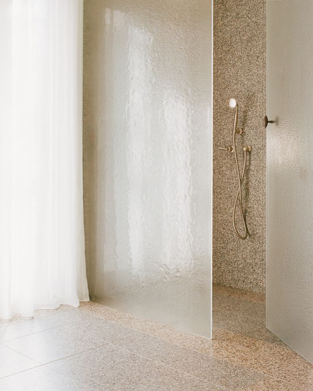 A minimalist walk-in bathroom shower with a frameless glass partition, textured pale plaster walls, and terrazzo flooring continuing into the shower area. A brass-gold handheld shower set is mounted against a speckled terrazzo feature wall, with a sheer white curtain at the side.
