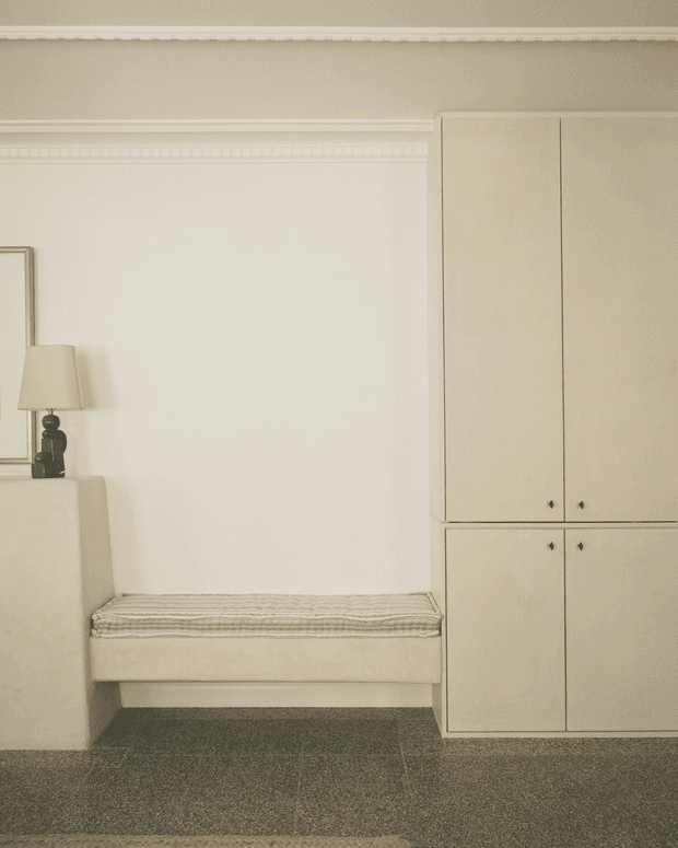A quiet, warmly lit traditional interior corner featuring a built-in upholstered bench seat with a striped cushion, tall cream built-in cabinetry, decorative wall moulding, a small table lamp, and dark textured carpet. The restrained palette and classic detailing create a refined, calm atmosphere.