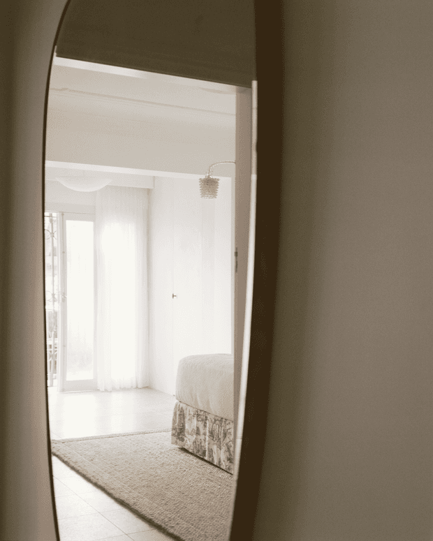 A softly lit bedroom viewed through a tall arched-edge mirror or doorway. The room features a pale upholstered bed with a patterned bed base, sheer floor-to-ceiling curtains, light tiled flooring, a neutral textured rug, white walls, and a delicate pendant light. The overall composition is minimalist, airy, and monochromatic.