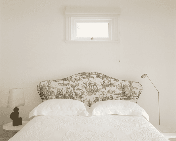 A softly lit, predominantly white bedroom with a neatly made bed featuring an ornate toile-patterned upholstered headboard, white embroidered bedding, a small round bedside table with a lamp and decorative object, and a slim wall-mounted reading lamp. A small high window with traditional white trim is centered above the bed.