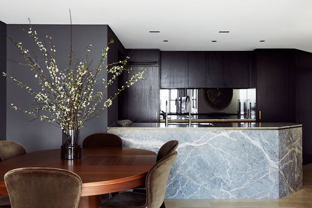 A sophisticated open-plan kitchen and dining area with dark timber cabinetry, a dramatic veined stone kitchen island, and a round rich-wood dining table with upholstered chairs. Integrated appliances and a reflective backsplash sit beneath a clean white ceiling with recessed lighting. The palette is dark, warm, and luxurious, with a large branch arrangement adding an organic element.