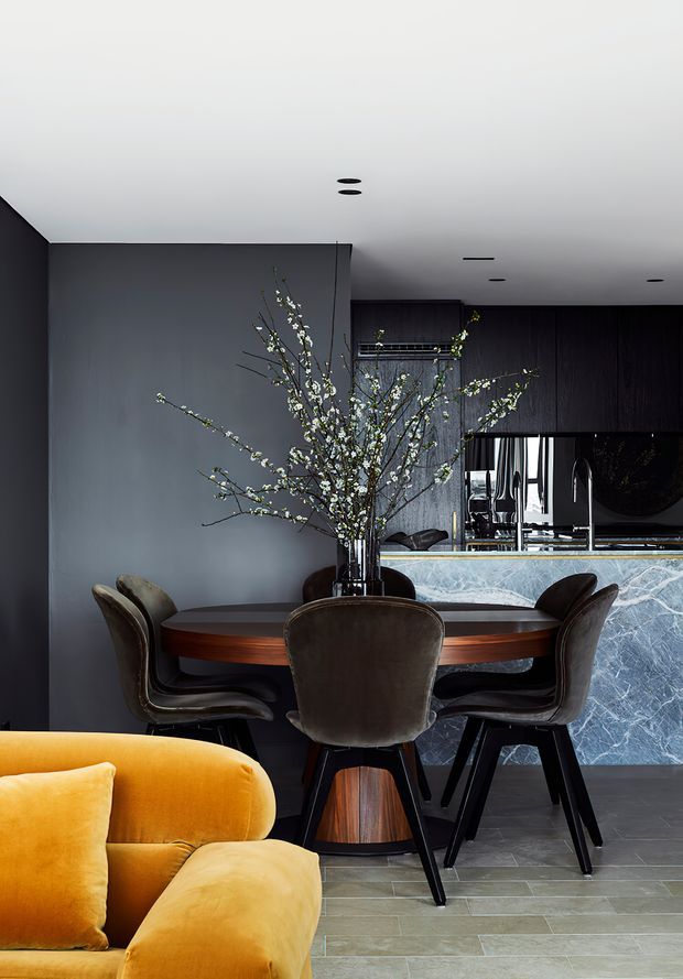 A moody, sophisticated open-plan dining and kitchen interior. A round dark timber dining table with upholstered brown chairs sits in front of a dark grey wall, while a dramatic veined stone kitchen island and dark timber cabinetry occupy the background. A bright mustard-yellow sofa adds a bold accent in the foreground, with a tall branch arrangement as a central decorative feature.