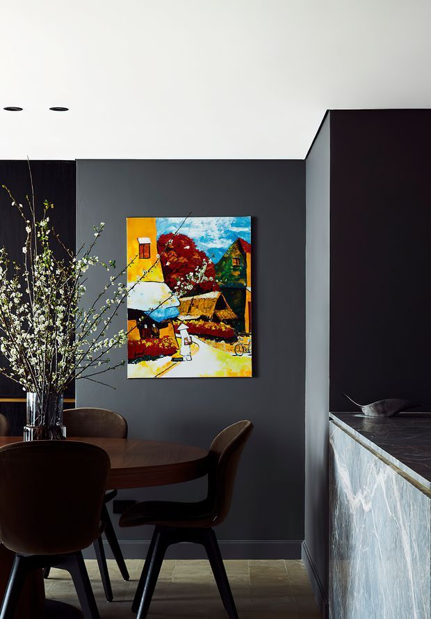 A refined dining area with a round dark timber dining table and sculptural dark chairs, set against charcoal-grey walls. A colourful framed artwork provides a vivid focal point, while a tall branch arrangement adds an organic element. To the right is a dark stone-topped island or counter with a textured pale stone-clad side, suggesting an adjoining kitchen. Recessed ceiling lights and restrained dark finishes create a dramatic, sophisticated interior.