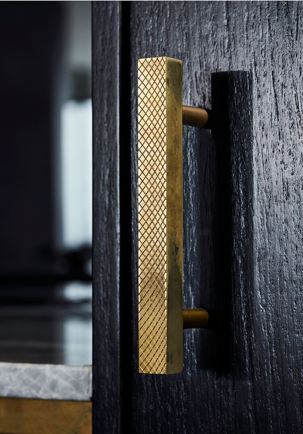 Close-up architectural detail of a tall brass-gold door pull with a diamond-textured face, mounted on a dark vertically textured door or wall panel. A dim interior and pale stone threshold are softly visible in the background.