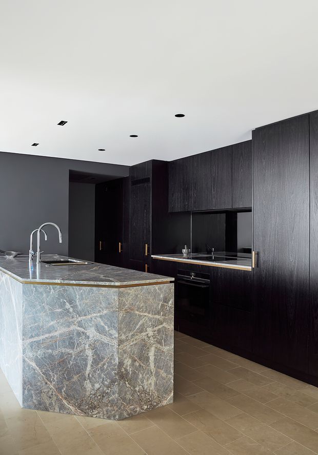 Contemporary dark-toned kitchen with full-height black timber-look cabinetry, integrated appliances, a large veined natural-stone island with waterfall edge, and a polished stone worktop. A double gooseneck tap and undermount sink sit at the island, while recessed ceiling lights and warm neutral floor tiles complete the refined scheme.