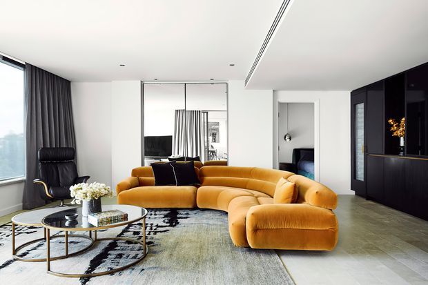 A refined contemporary living room with a large curved mustard velvet sectional sofa, black leather lounge chair, round glass coffee tables, and a neutral patterned rug. The room has white walls and ceiling, floor-to-ceiling glazing with dark curtains, mirrored sliding panels, polished grey flooring, and a dark built-in entertainment or bar joinery wall. A bedroom is visible through an open doorway in the background.