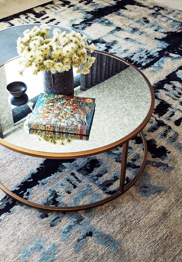 Close-up interior vignette featuring nested round coffee tables with reflective glass tops and warm timber-toned frames, placed over a large abstract patterned rug in cream, taupe, black, and blue. The tables hold a dark textured vase of pale flowers, a small dark bowl, and a colourful art book. A dark ribbed architectural or furniture element is reflected in the glass.