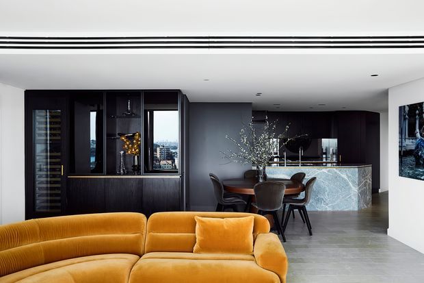 Contemporary open-plan living and dining area with a dramatic mustard velvet sectional in the foreground, dark timber built-in cabinetry and wine storage along the left wall, a round dining table with upholstered chairs, and a marble-clad kitchen island or bar at the rear. The space has a restrained palette of charcoal, grey, white, and warm ochre, with recessed ceiling lighting and city views through the windows.