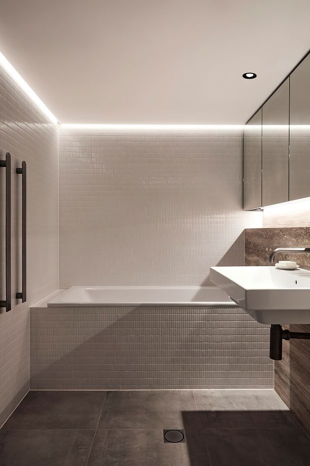 A contemporary compact bathroom featuring a wall-to-wall bathtub, white vertically textured mosaic wall tiles, dark large-format floor tiles, a floating white basin, dark exposed plumbing, a stone-look vanity wall, frameless glass shower screening, and integrated linear LED lighting around the ceiling perimeter. The space has a refined, warm and softly dramatic atmosphere.