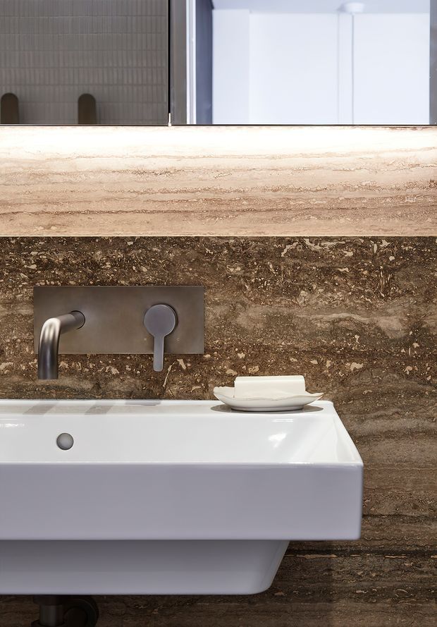 Close-up view of a refined contemporary bathroom vanity with a wall-mounted white ceramic basin, brushed dark-metal wall-mounted tapware, and a small integrated overflow. The basin is set against richly veined brown natural stone wall cladding, with a pale horizontal stone ledge and a mirror above. The composition highlights the tactile stone surfaces and minimalist bathroom fixtures.