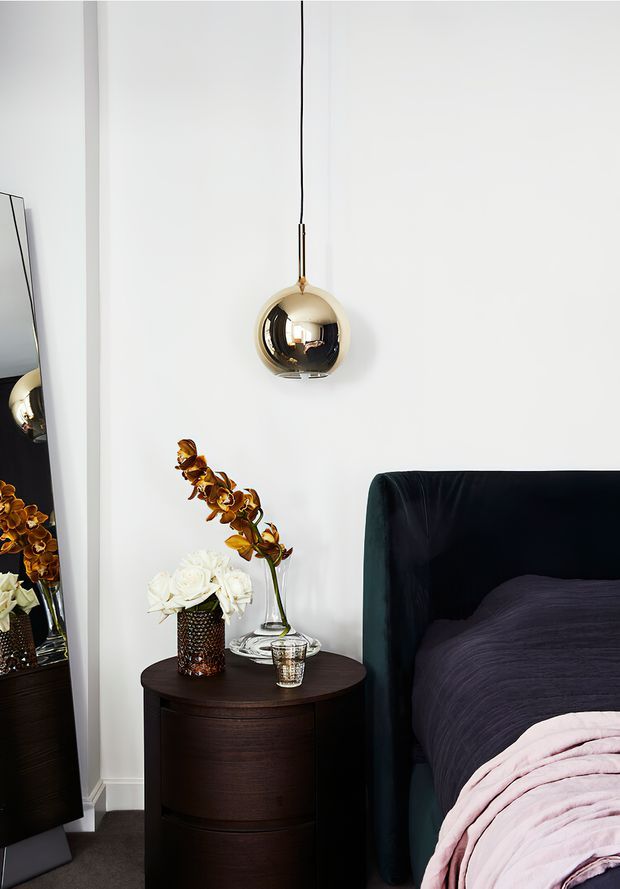 A refined bedroom vignette featuring a dark upholstered bed with a deep purple duvet and soft pink throw, a rounded dark wood bedside table, white and amber orchid arrangements, and a spherical brass pendant light suspended against a white wall. A tall mirror reflects the bedside styling on the left.