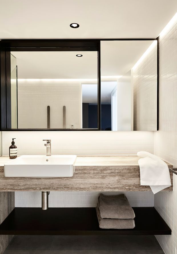 Contemporary bathroom vanity with a large black-framed mirror, white vessel basin, brushed metal tapware, and a thick textured stone benchtop. White vertically textured wall tiles, concealed linear lighting, and dark open shelving create a refined, high-contrast minimalist interior.