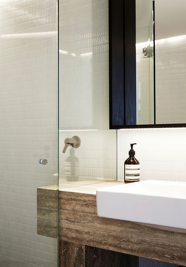 A close-up contemporary bathroom vanity with a white rectangular basin mounted on a textured brown stone-look vanity. The shower area is enclosed by clear glass, with small white vertical tiles covering the walls. A dark timber-framed mirrored cabinet with integrated underlighting hangs above the vanity, and brushed metal tapware and a dark soap dispenser provide subtle contrast.
