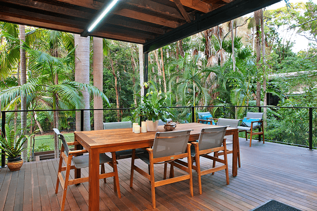 A covered outdoor dining deck surrounded by lush tropical vegetation. A solid timber dining table with six timber-framed chairs occupies the centre, with potted plants and simple table decor. The deck has dark timber floorboards, exposed timber roof framing, slim dark balustrades, and a compact linear light under the roof.
