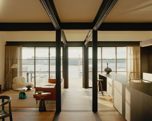 A warm, sophisticated open-plan interior with strong black structural framing dividing the living, dining, and kitchen areas. Full-height glazed doors open to a balcony overlooking water. The living area contains curved upholstered seating and a floor lamp, while the kitchen features a long pale island with integrated seating and dark bar stools. Timber flooring, neutral walls, and abundant natural light create a refined mid-century-inspired atmosphere.