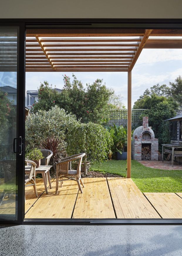 View from inside through large dark-framed sliding glass doors onto a timber deck and landscaped backyard. The deck has a timber pergola with open rafters, outdoor wicker seating, dense garden planting, and a freestanding brick outdoor pizza oven with a small adjacent timber structure. A terrazzo-like interior floor is visible in the foreground.