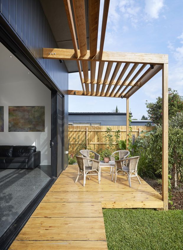 A contemporary outdoor timber deck and entertaining area beside a house, featuring a large exposed-timber pergola with overhead rafters, wicker dining chairs around a small table, garden planting, and a black-framed glazed opening into the living room. The space is bright and informal, with strong indoor-outdoor connection.