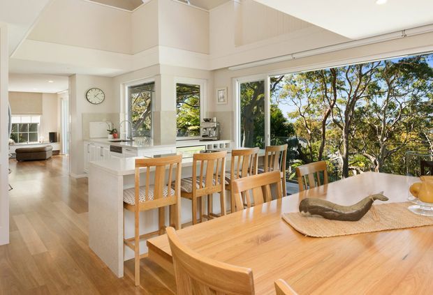 Bright open-plan residential kitchen and dining area with a white island, integrated kitchen cabinetry, timber flooring, and a large timber dining table with matching chairs. Floor-to-ceiling glazing and sliding doors open to leafy outdoor views, while a partial living room is visible beyond the kitchen. The space has high ceilings, abundant natural light, and a warm neutral palette.