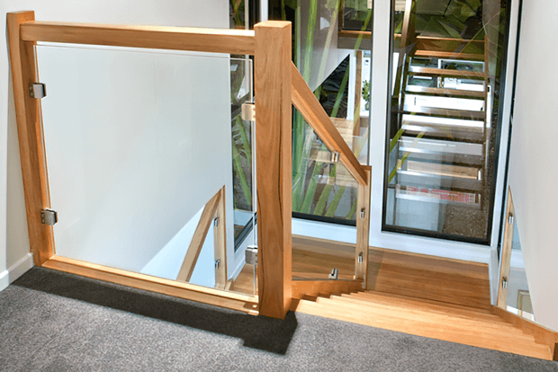 Interior view of a stairway and landing featuring a warm timber staircase, carpeted upper landing, clear glass balustrade panels, and black-framed floor-to-ceiling glazing. The stairs descend beside a bright open void with a matching staircase visible through the glazing.