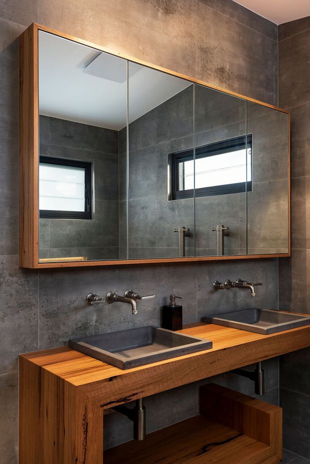 Contemporary bathroom vanity with a double timber-framed mirror cabinet, two dark rectangular vessel basins, wall-mounted metal taps, and a thick timber countertop with open shelving below. Large dark grey concrete-look wall tiles create a moody, industrial-inspired backdrop, while the mirrors reflect black-framed windows and the ceiling.