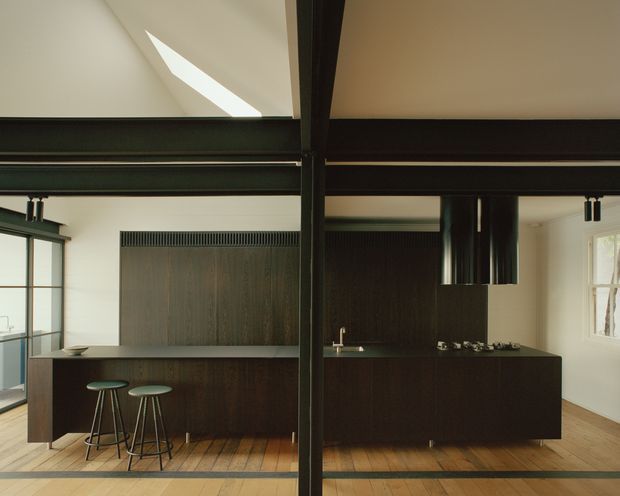 A contemporary open-plan kitchen viewed straight on, featuring dark timber cabinetry and island units, black exposed structural beams, a black rangehood, integrated cooktop and sink, timber flooring, and two bar stools. White walls, clerestory-like glazing, and a skylight bring natural light into the space.