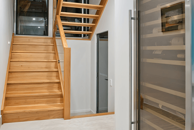 Interior view of a contemporary stairway and landing with warm timber stairs, open timber balustrade, white walls, and a glass-fronted storage or display area. A doorway opens beneath the stairs, while glazing and reflected shelving add depth to the compact circulation space.