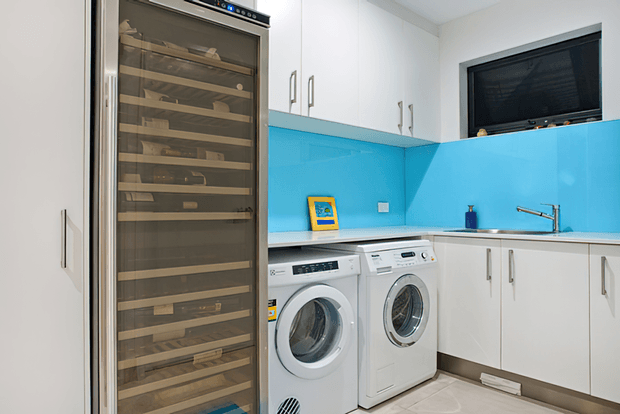 Bright, compact contemporary laundry room with white overhead and base cabinetry, a vivid turquoise-blue splashback, integrated front-loading washing machine and dryer, a stainless sink with tap, and a tall glass-fronted wine refrigerator or storage unit. A wall-mounted television is recessed above the cabinetry.