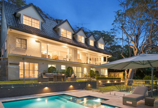 Twilight exterior view of a substantial multi-storey residence with a dark tiled pitched roof, repeated dormer windows, pale masonry walls, extensive illuminated upper-level glazing and a covered terrace. The landscaped rear yard includes a swimming pool, raised planting beds, outdoor seating, umbrellas, steps and integrated landscape lighting, with mature trees surrounding the property.