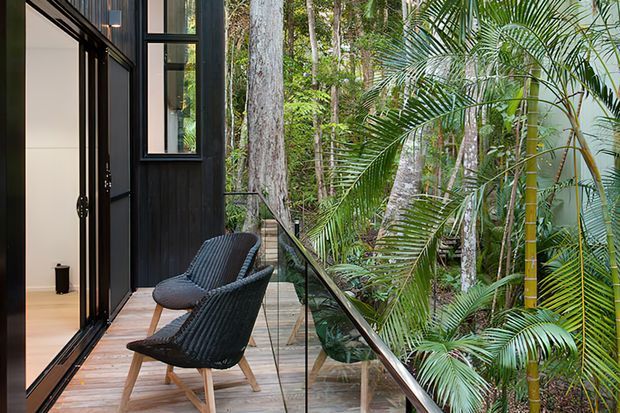 A compact timber balcony or terrace beside a dark-clad modern building, furnished with two woven lounge chairs. Full-height black-framed glazing opens onto the deck, while a clear glass balustrade overlooks dense tropical vegetation and tall trees.