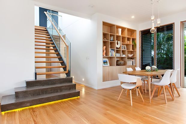 Bright contemporary open-plan dining and stair area with a custom timber built-in display shelving unit, round timber dining table, white chairs, and a floating timber staircase with glass balustrade. Pale timber flooring, white walls, warm recessed lighting, and large windows create an airy, refined interior.