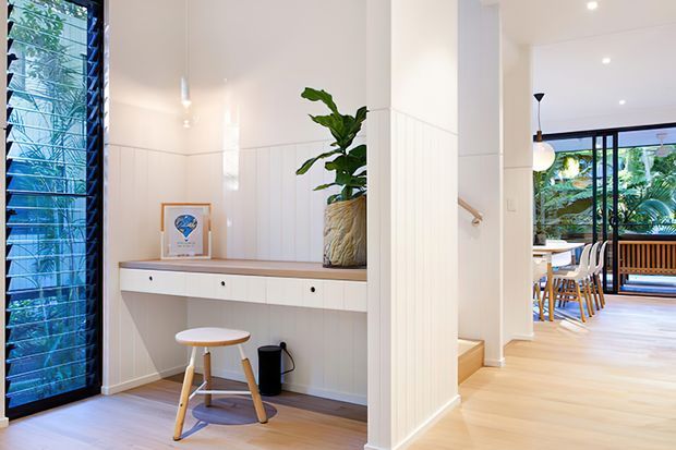 Bright, contemporary residential entry and hallway with white vertical wall panelling, pale timber flooring, a built-in console and stool, indoor plant, and a glazed louvre door. The hallway opens toward a dining area with timber furniture and abundant garden views through large windows.
