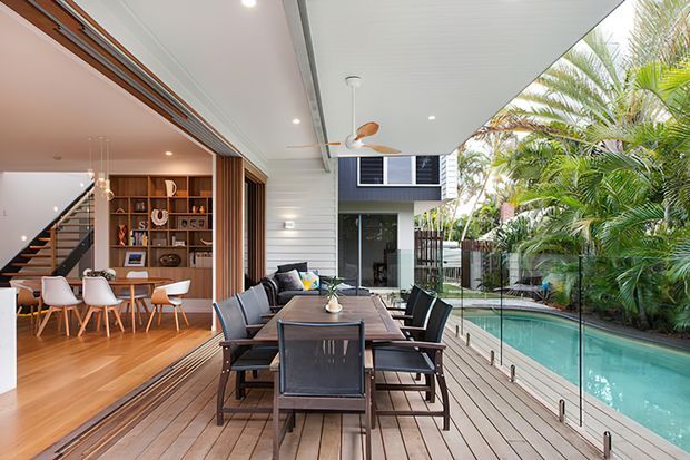 A bright, contemporary indoor-outdoor dining area on a timber deck beside a swimming pool. The covered terrace has a large timber dining table with dark chairs, a ceiling fan, and frameless glass balustrades. Through wide open glazing, a warm timber-floored living area with built-in shelving, staircase, and vertical timber screening is visible. Tropical planting borders the pool.
