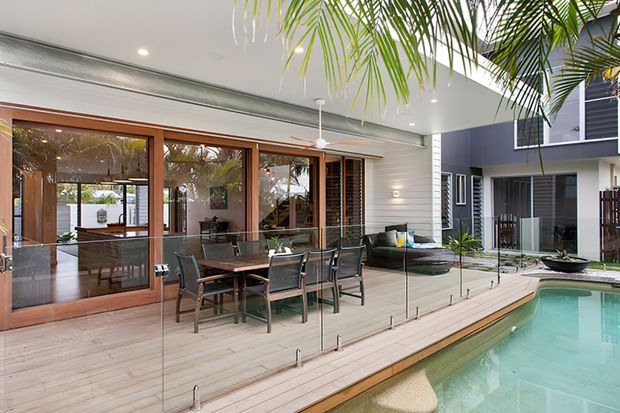 Covered outdoor entertaining area beside a swimming pool, with timber decking, frameless glass pool fencing, a large dining table, and lounge seating. The space connects to the interior through timber-framed glazed doors and features white horizontal wall cladding, recessed lighting, and tropical palm planting.