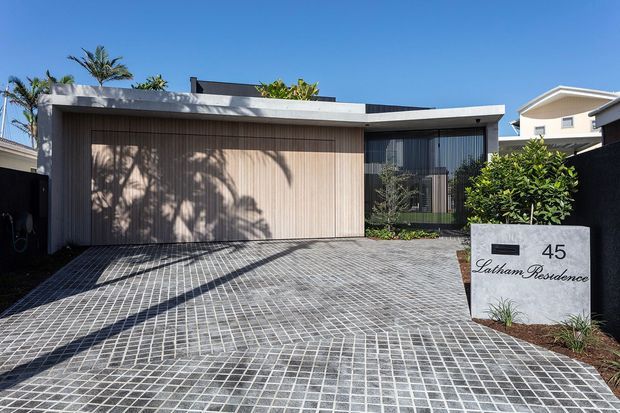 Street-facing view of a contemporary single-storey residence with a flat roof, broad timber-look garage door and dark-framed glazing. The foreground is a paved driveway with a grid-like pattern, while landscaped planting and a residential address sign sit beside the entry.