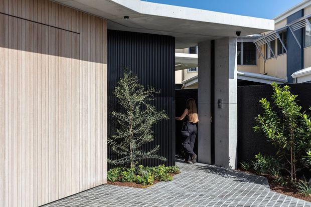 A contemporary residential entrance viewed from outside, featuring a cantilevered flat concrete canopy, a tall concrete entry portal, vertical light timber cladding, a black ribbed feature wall, and a dark pivot door. The approach is paved with small grey grid-pattern pavers and framed by planted garden beds. A neighbouring multi-storey residential building is visible in the background.