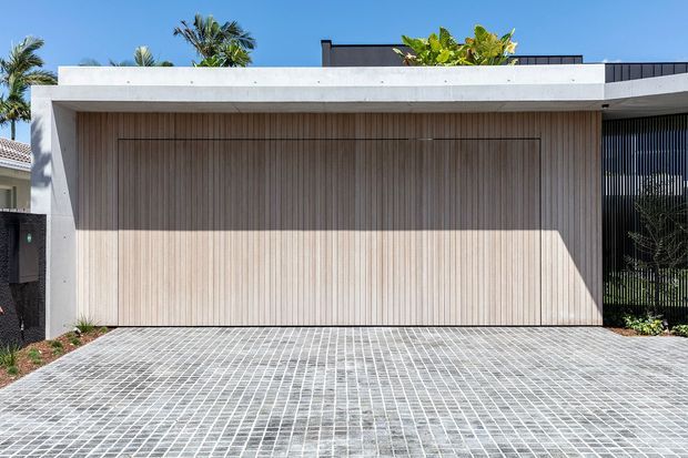 A contemporary residential facade viewed from the driveway, featuring a large timber-look garage door set within a smooth white rendered portal. The flat roofline and dark vertical screening create a clean modern composition, with pale grid-patterned paving in the foreground and tropical planting visible around the edges.