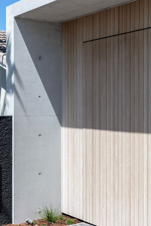 Close-up exterior view of a contemporary garage or entry door framed by a smooth raw concrete surround. The tall door has vertically grooved light timber cladding with a slim dark horizontal reveal, while strong sunlight casts geometric shadows across the concrete and timber surfaces. A small area of landscaping and dark textured wall cladding is visible at the base and side.
