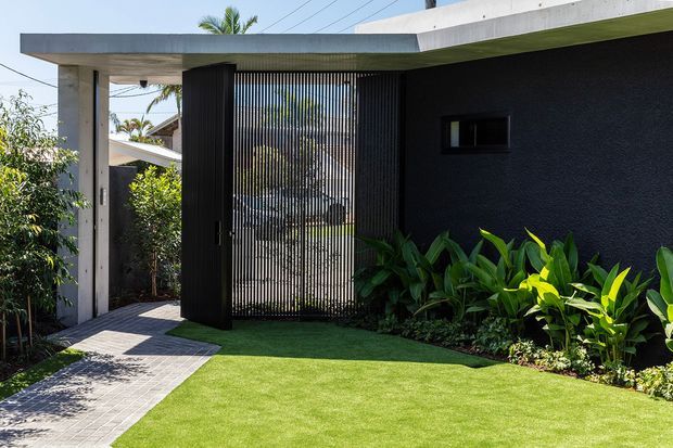 A contemporary single-storey home entry viewed from the landscaped front yard. The facade combines a flat concrete roof slab, dark textured wall finish, exposed concrete columns, and a tall black vertical screening gate. A paved walkway crosses a manicured lawn, with tropical planting along the wall.
