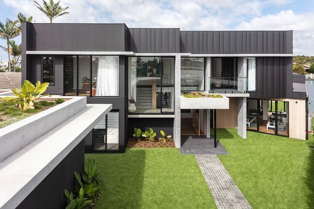 A contemporary multi-level residential home with a bold rectilinear form, dark vertical metal cladding, concrete terraces and projecting balcony elements. Large glazed openings reveal interior living spaces, while landscaped lawn, raised planters and a tiled pathway surround the building. Tropical planting and bright daylight contribute to a modern suburban setting.