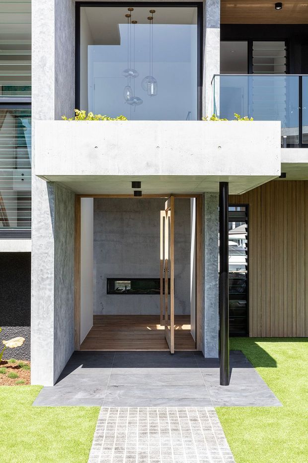 Contemporary multi-storey residential facade viewed from the front, featuring a recessed entry beneath a projecting concrete balcony. The entrance has a tall timber pivot door, dark tiled paving, exposed concrete walls and soffit, a black slender structural post, timber-look vertical cladding, glass balcony balustrading, and large glazed openings. A manicured lawn and tiled path lead to the entry.