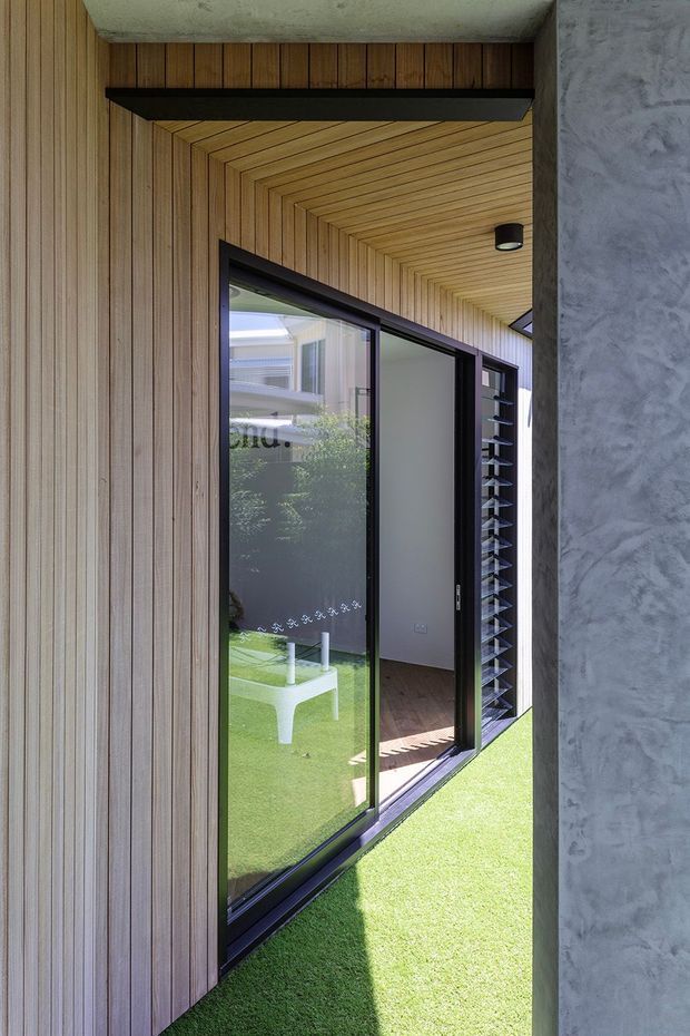 A narrow, sheltered entry or passage with vertical timber wall panelling and a timber-lined ceiling. Black-framed glazed sliding doors open toward a small lawn, with a louvred window panel beyond. A textured grey concrete or render wall forms the right side, and a black structural beam and compact ceiling light add contrast. The image has a modern, material-focused architectural character with strong natural light and indoor-outdoor connection.